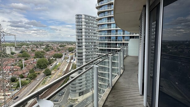 A modern building with a curved design, featuring a balcony with a metal railing and a view of a cityscape with residential areas and a railway track. - Charming 2-bed Apartment in London (London)