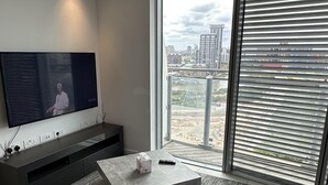 A modern living room with a flat-screen TV, a grey sofa, a marble coffee table, and a large window with blinds. - Charming 2-bed Apartment in London (London)