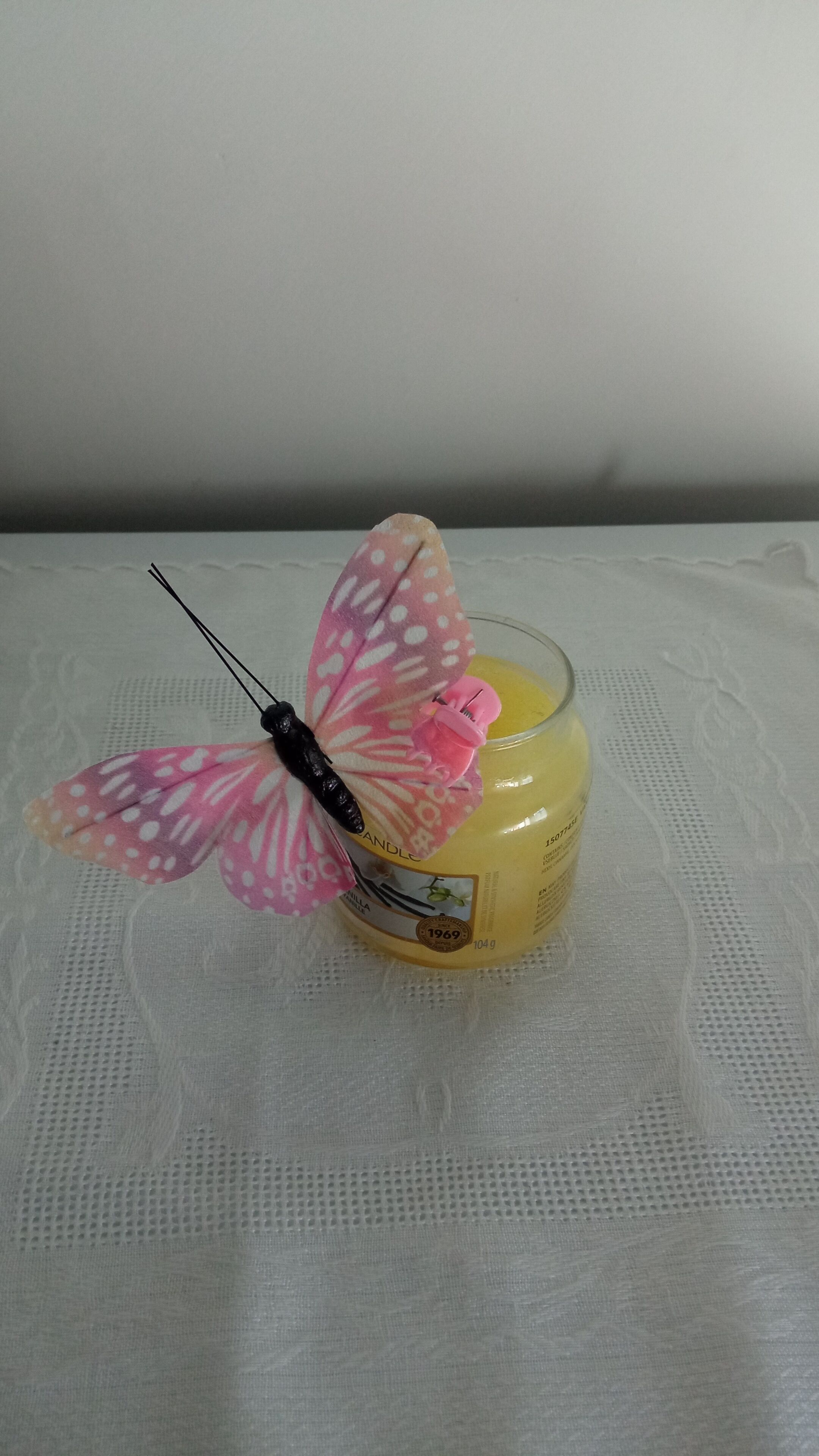 A candle with a pink butterfly decoration on a lace tablecloth.