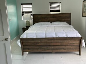 A bedroom with a wooden bed, two bedside tables, a window with blinds, and a door.