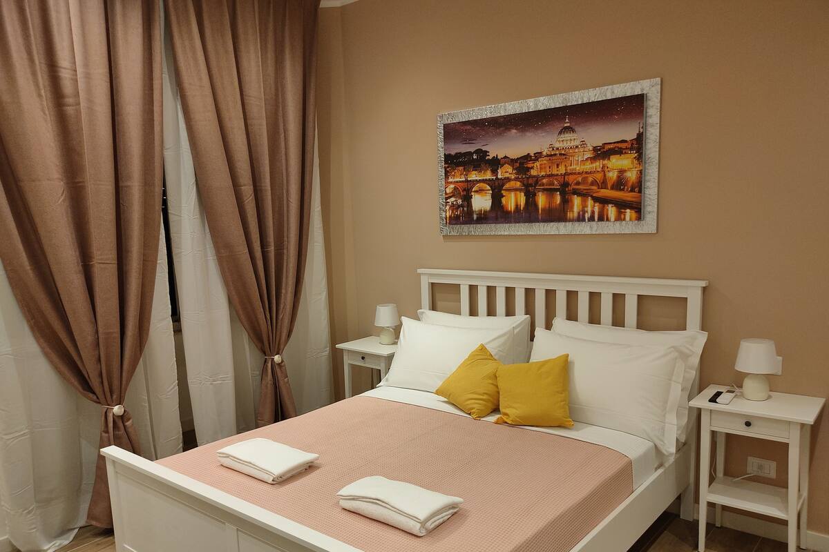 Una cama con arreglos impecables, con almohadas blancas y amarillas, una colcha rosa y toallas blancas dobladas sobre la cama. Hay una mesita de noche blanca con una lámpara y un teléfono encima. Un cuadro de un paisaje urbano adorna la pared sobre la cama.