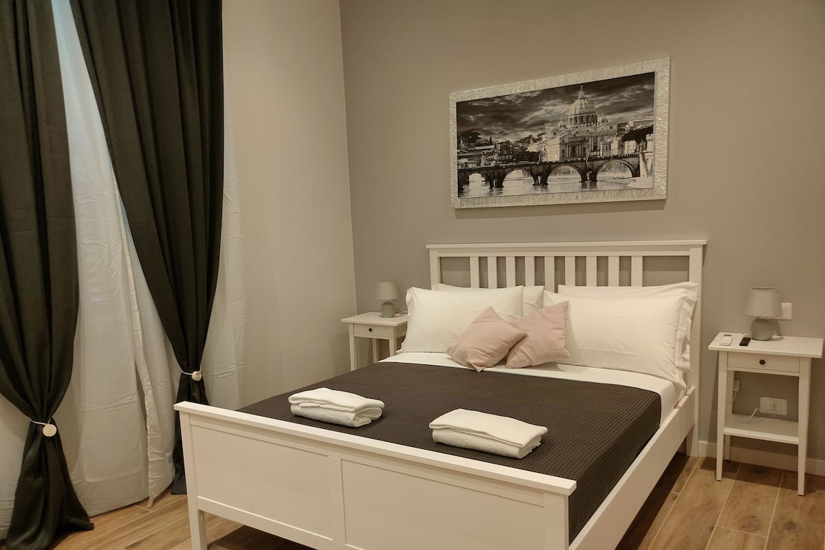 Una cama bien hecha con ropa de cama blanca y dos almohadas, una mesita de noche con una lámpara, un cuadro colgado en la pared y una cortina a la izquierda.