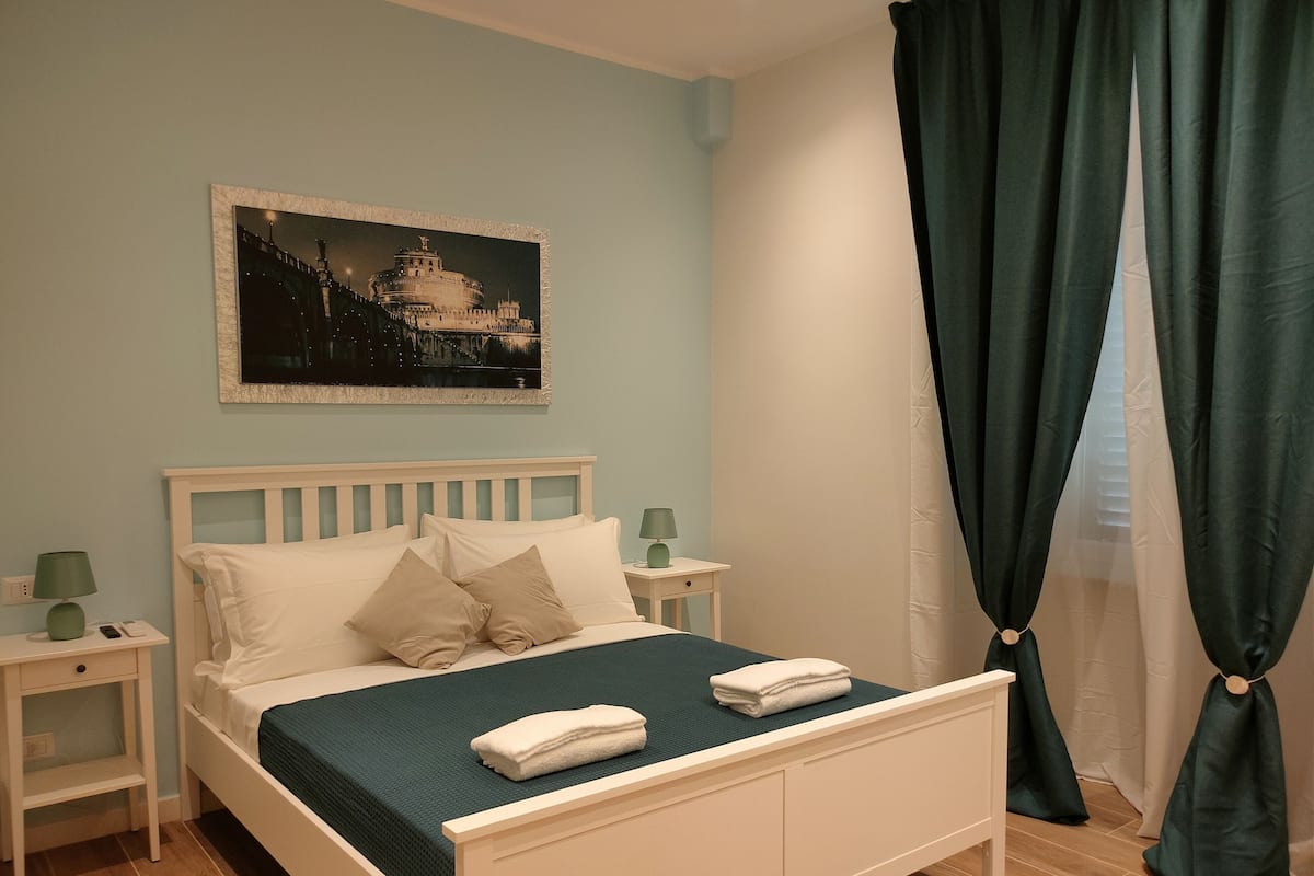 Una cama bien tendida con ropa de cama y almohadas blancas, un edredón verde y una toalla doblada en la cabecera. Hay una mesita de noche con una lámpara y un cuadro enmarcado en la pared sobre la cama.