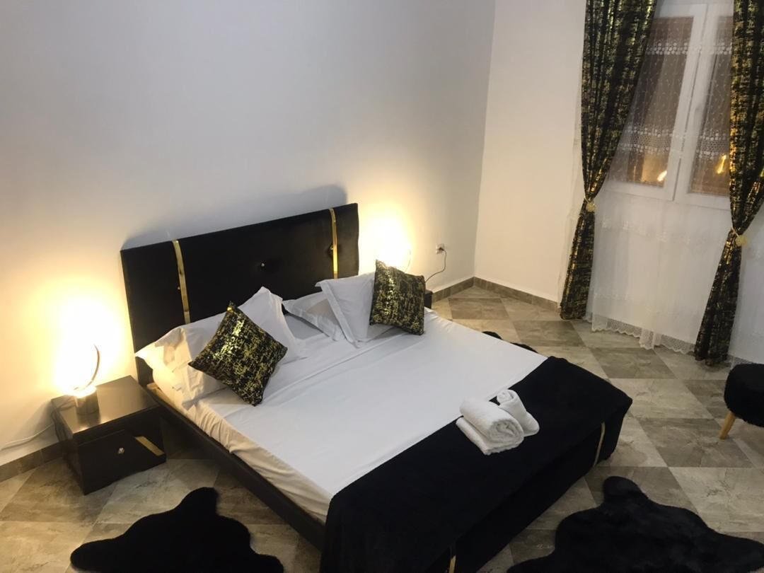 Een net opgemaakt bed met witte lakens en twee decoratieve kussens, een nachtkastje met een lamp, en een raam met gordijnen.