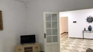 A room with a television on a wooden stand, a white door, a tiled floor, and a small table with decorative items.