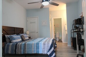 Une chambre à coucher avec un lit, un ventilateur de plafond, une porte et une salle de bain visible au fond.