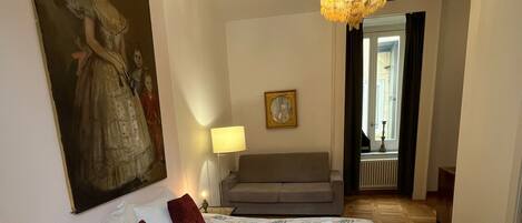 A bedroom with a large bed, a painting on the wall, a chandelier, and a view of the outside.