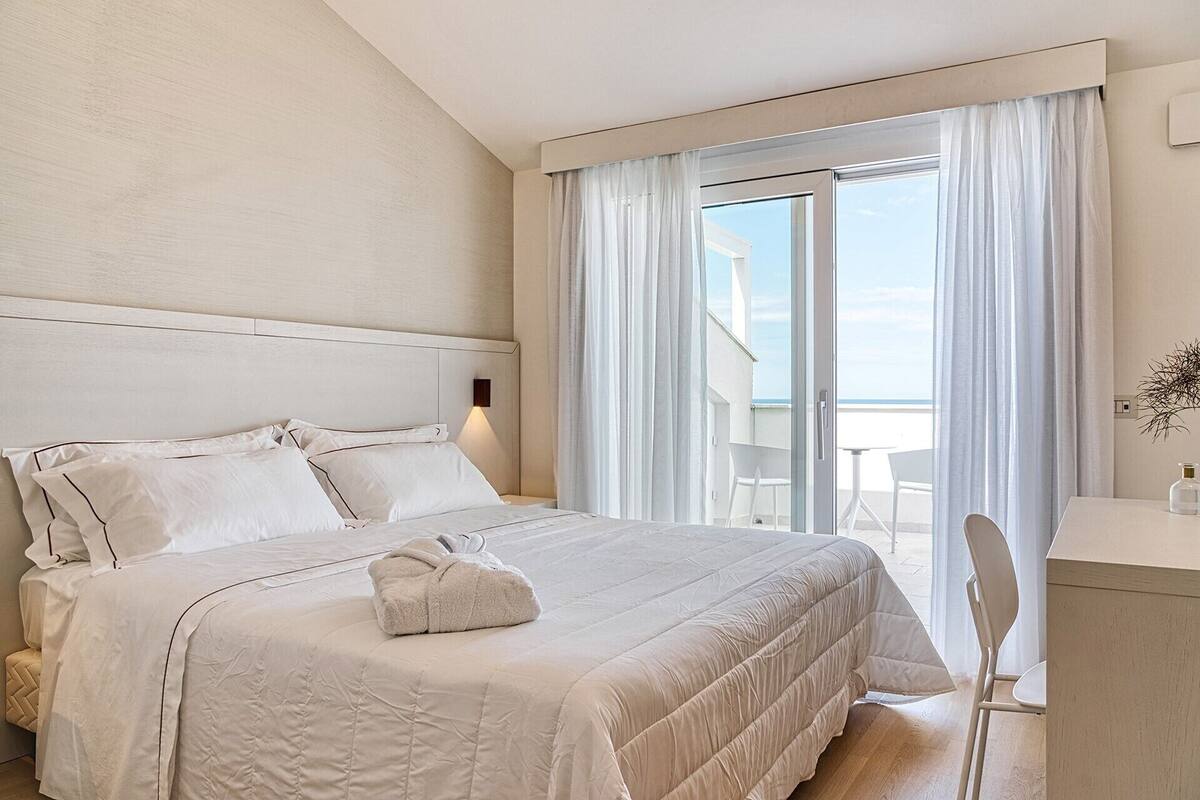 Hotellihuone, jossa on sänky, yöpöytä, tuoli ja suuri ikkuna, josta avautuu näkymä merelle.