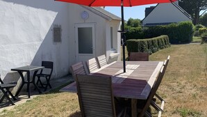 An outdoor dining area with a wooden table and chairs, a red umbrella, and a white building in the background.