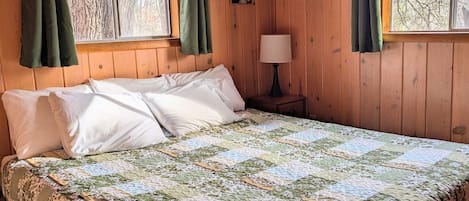 Une chambre dans une cabane en bois, avec un grand lit, des rideaux verts, un ventilateur de plafond et un tableau encadré au mur.