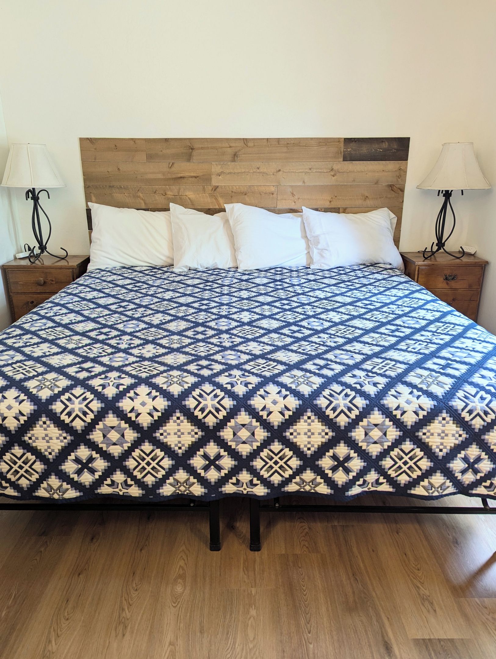 Una cama con una colcha estampada, cabecera de madera, dos mesitas de noche con lámparas y suelo de madera.