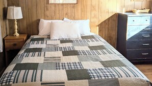 A bedroom with a wooden paneled wall, a bed with a quilt, a nightstand with a lamp, and a dresser. - Elms Cabin at Lazy Oaks Resort (Lakeside)