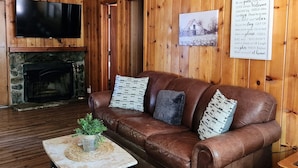 Un salon chaleureux avec un canapé en cuir, un téléviseur à écran plat, une cheminée en pierre et un mur en bois orné d’une inscription de bienvenue.