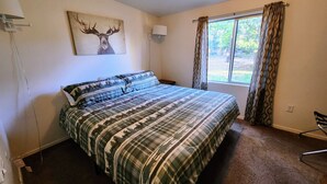 A bedroom with a large bed, a window with curtains, a wall-mounted lamp, and a deer painting. - Pines Cabin at The Lazy Oaks Resort (Lakeside)