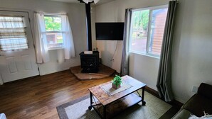 A living room with a wooden floor, a black wood-burning stove, a ceiling fan, a flat-screen TV, a wooden coffee table, and a green plant. - Pines Cabin at The Lazy Oaks Resort (Lakeside)