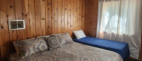 Una habitación con paredes de paneles de madera, una cama con ropa de cama estampada, una cama más pequeña con ropa de cama azul, una ventana con cortinas blancas y una rejilla de ventilación en el techo.