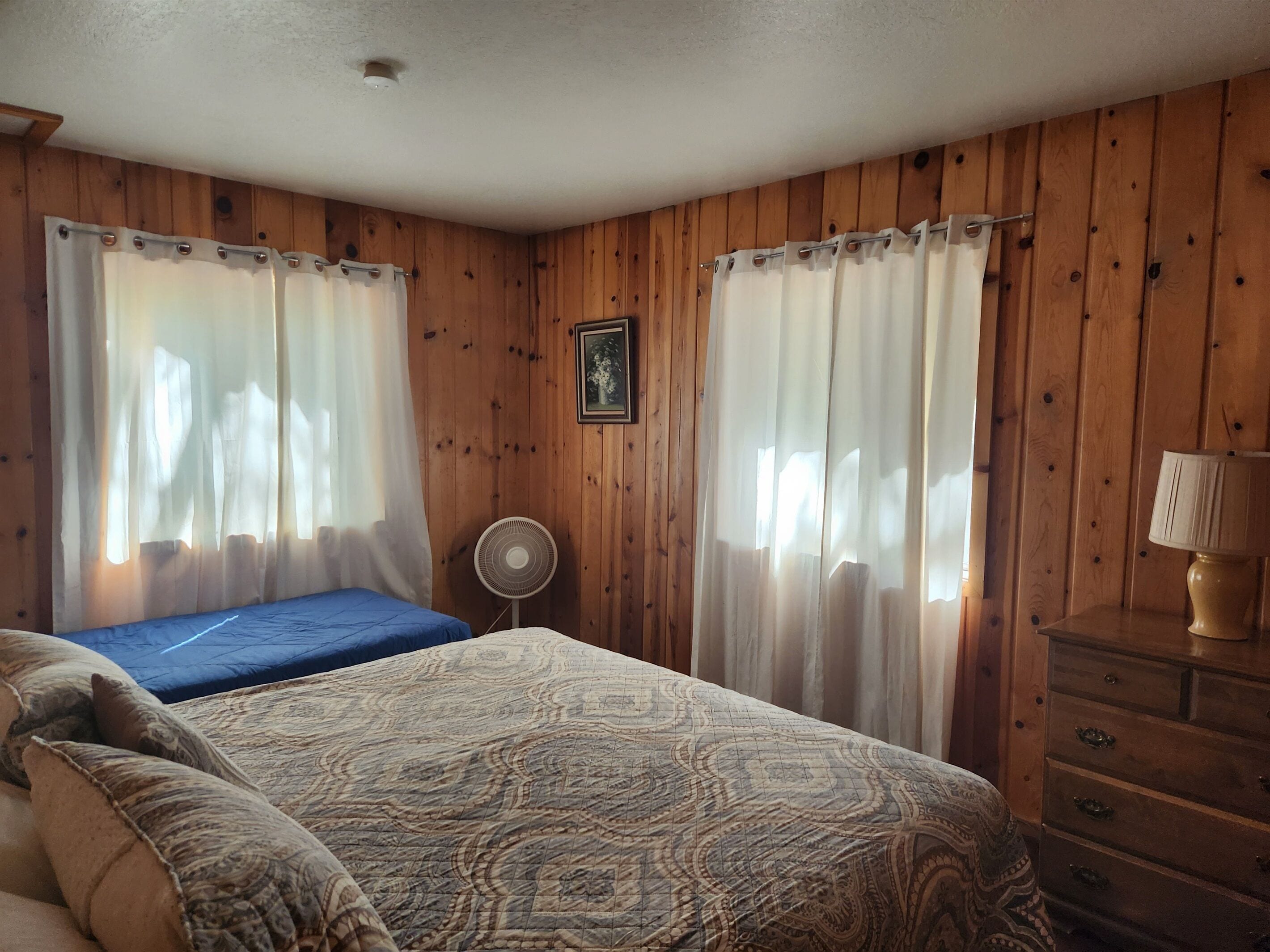 Una camera da letto con un letto, due finestre con tende leggere, una ventola sul comodino, una cassettiera e un quadro appeso al muro.