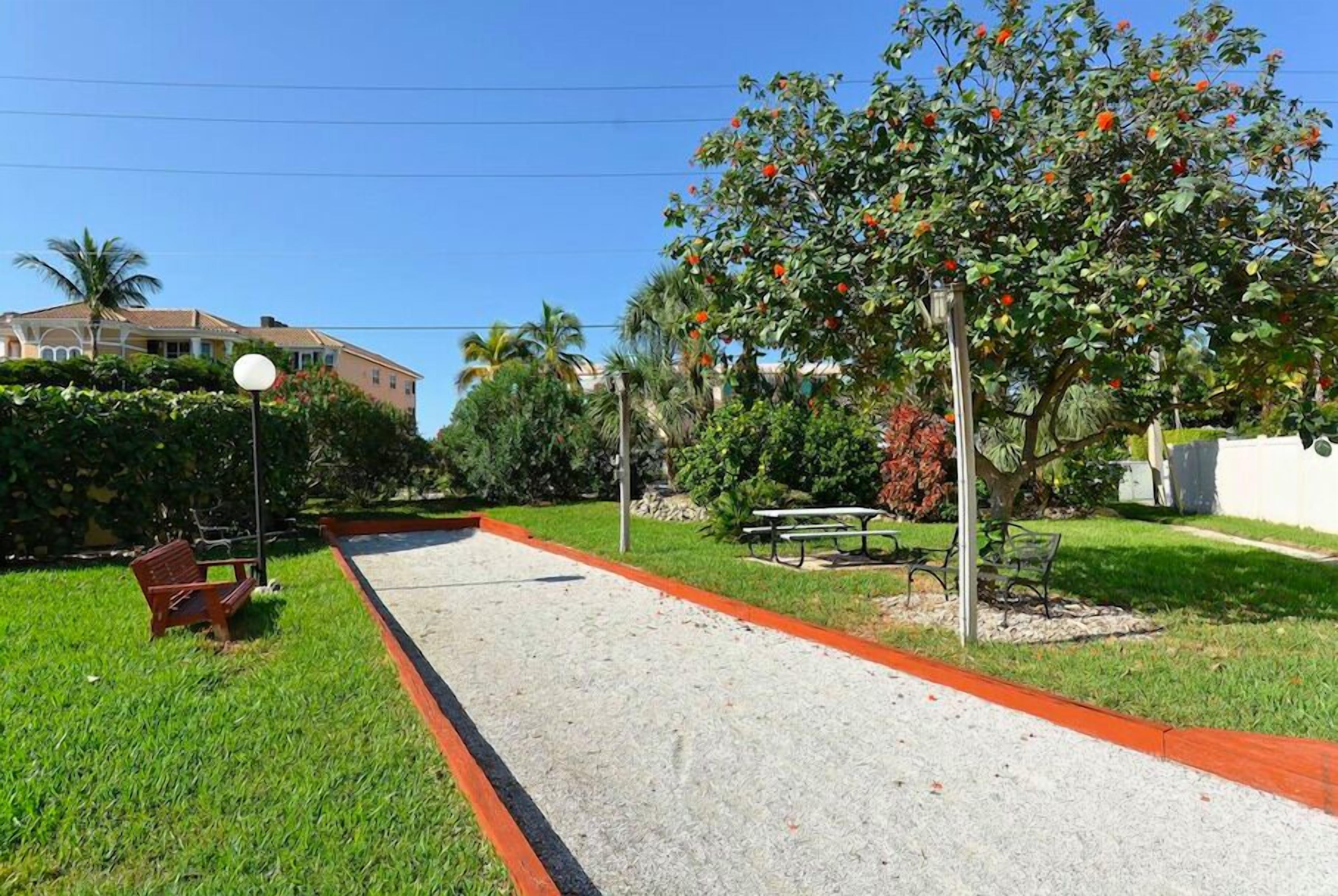 A paved walkway with a picnic table and benches, surrounded by greenery and a palm tree.