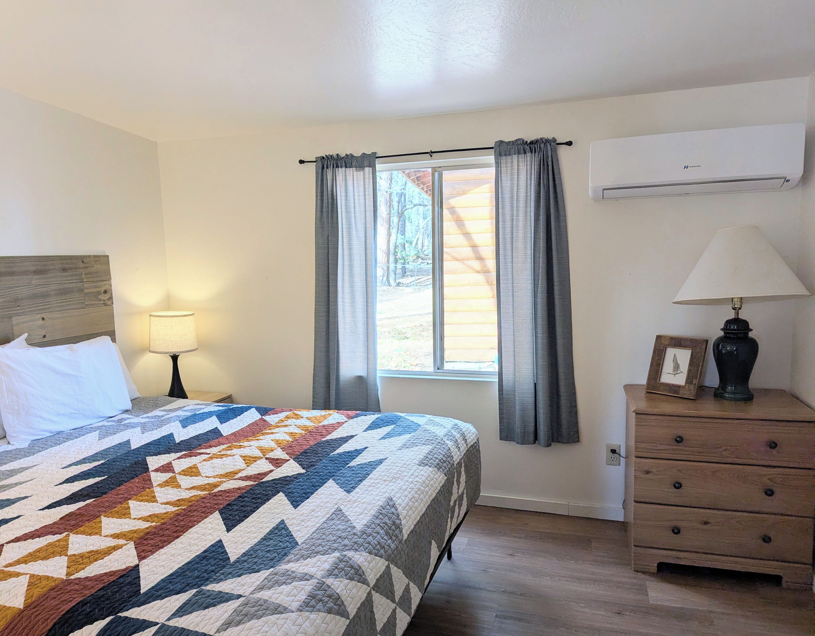 Un dormitorio con una cama, una mesita de noche, un aparador, una ventana con cortinas y un aire acondicionado.
