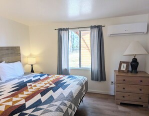 A bedroom with a bed, a nightstand, a dresser, a window with curtains, and an air conditioner.