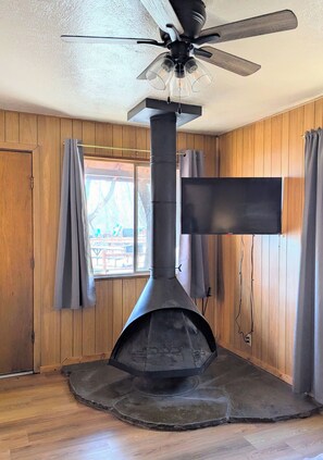 A room with a wood-paneled interior, a ceiling fan, a large black fireplace, and a window with curtains. - Willows Cabin at Lazy Oaks Resort (Lakeside)
