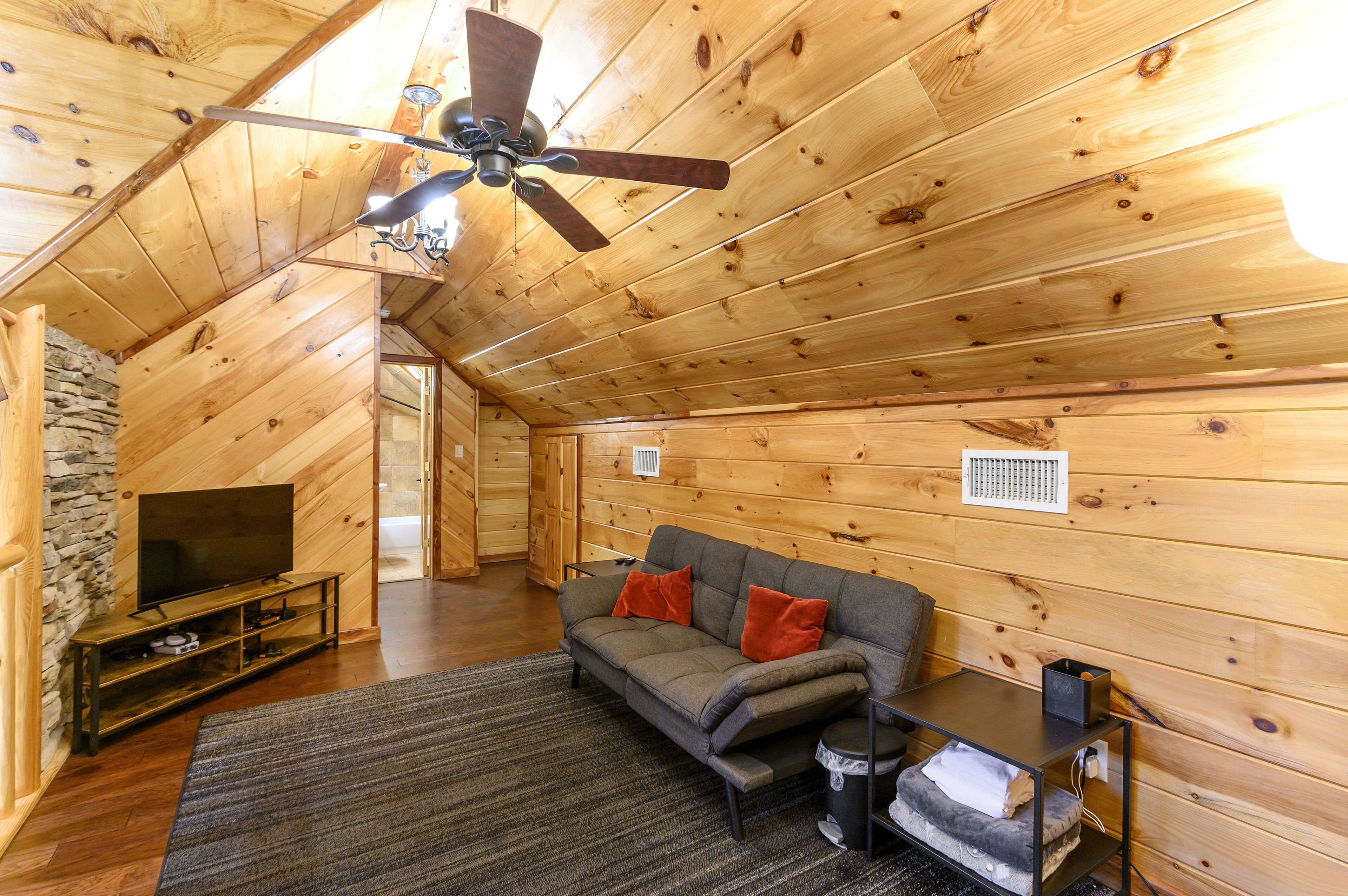 A log cabin interior with a wooden ceiling, a ceiling fan, a sofa with cushions, a television on a wooden stand, and a stone wall.