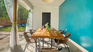 A wooden table with wicker chairs, a hanging light fixture, and a view of a backyard with a wooden fence and greenery. - Maldives Vibe | Ping-Pong | Firepit I Yoga (Austin)