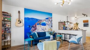 A living room with a large painting of a coastal scene, a blue sofa, a coffee table, and a guitar on the wall. - Santorini Inspired | Firepit | Grill I Yoga (Austin)