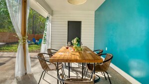 An outdoor dining area with a wooden table and wicker chairs, a hanging light fixture, and a view of the backyard. - Fun Group Travel Retreat (Austin)