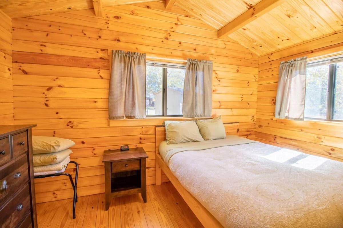 A wooden cabin bedroom with a bed, two windows with curtains, a nightstand, and a dresser.