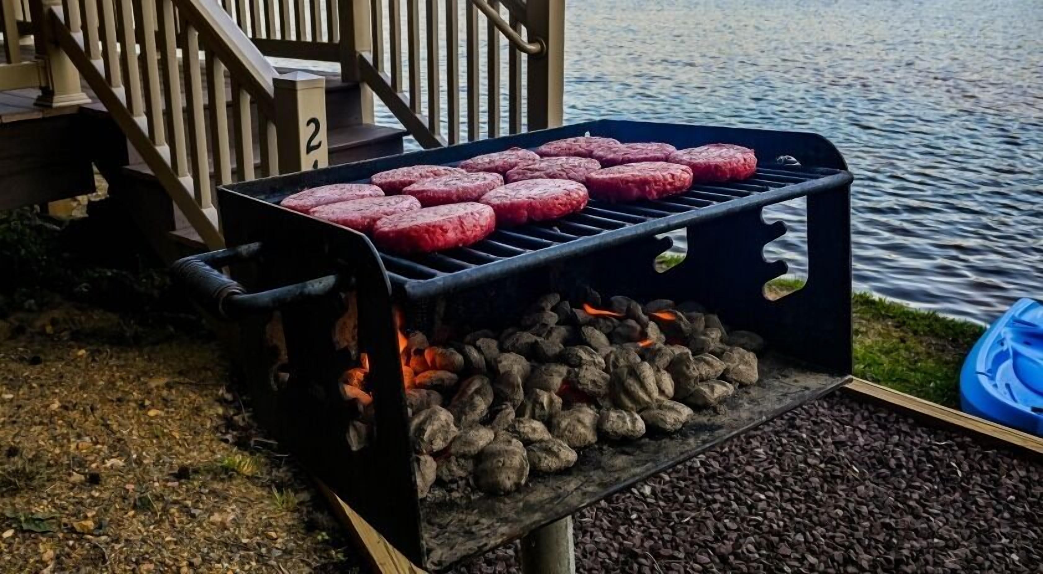 A grill with multiple burgers cooking over charcoal, set against a lakeside backdrop.