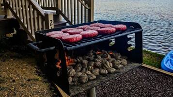 A grill with multiple burgers cooking over charcoal, set against a lakeside backdrop.