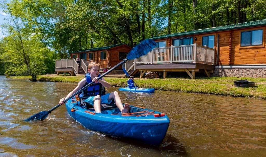A person in a blue kayak on a river, with a cabin in the background.