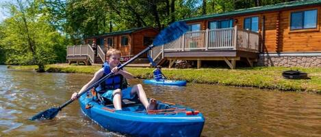 A person in a blue kayak on a river, with a cabin in the background.