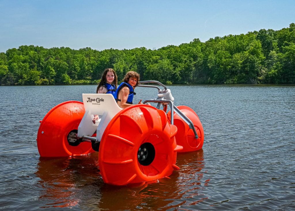 A red amphibious vehicle on a lake with two people inside, surrounded by a forested shoreline.