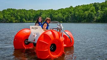 A red amphibious vehicle on a lake with two people inside, surrounded by a forested shoreline.
