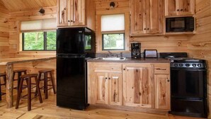 A wooden cabin interior with a kitchen featuring a black refrigerator, stove, and microwave, wooden cabinets, and a bar area with stools. - Wilderness Presidential Resort (Spotsylvania)