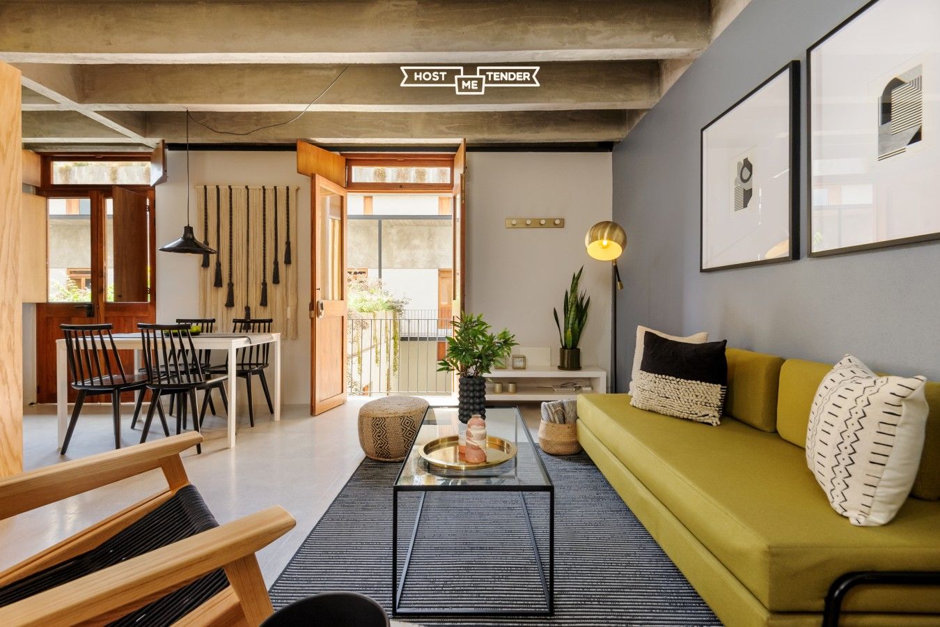A modern living room with a sofa, coffee table, and chairs. There is a dining area with a table and chairs. The room has a large window and a door leading outside.