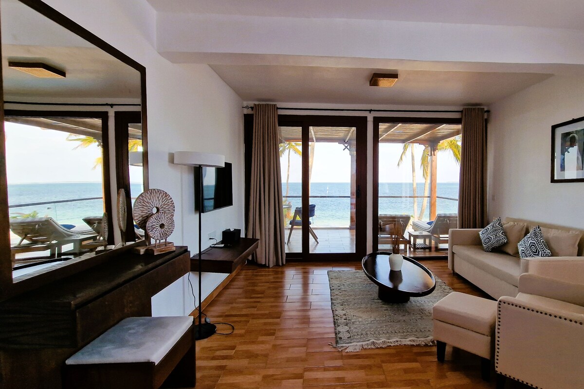 A living room with a sofa, coffee table, and TV, a wooden floor, and a view of the ocean through large glass doors.