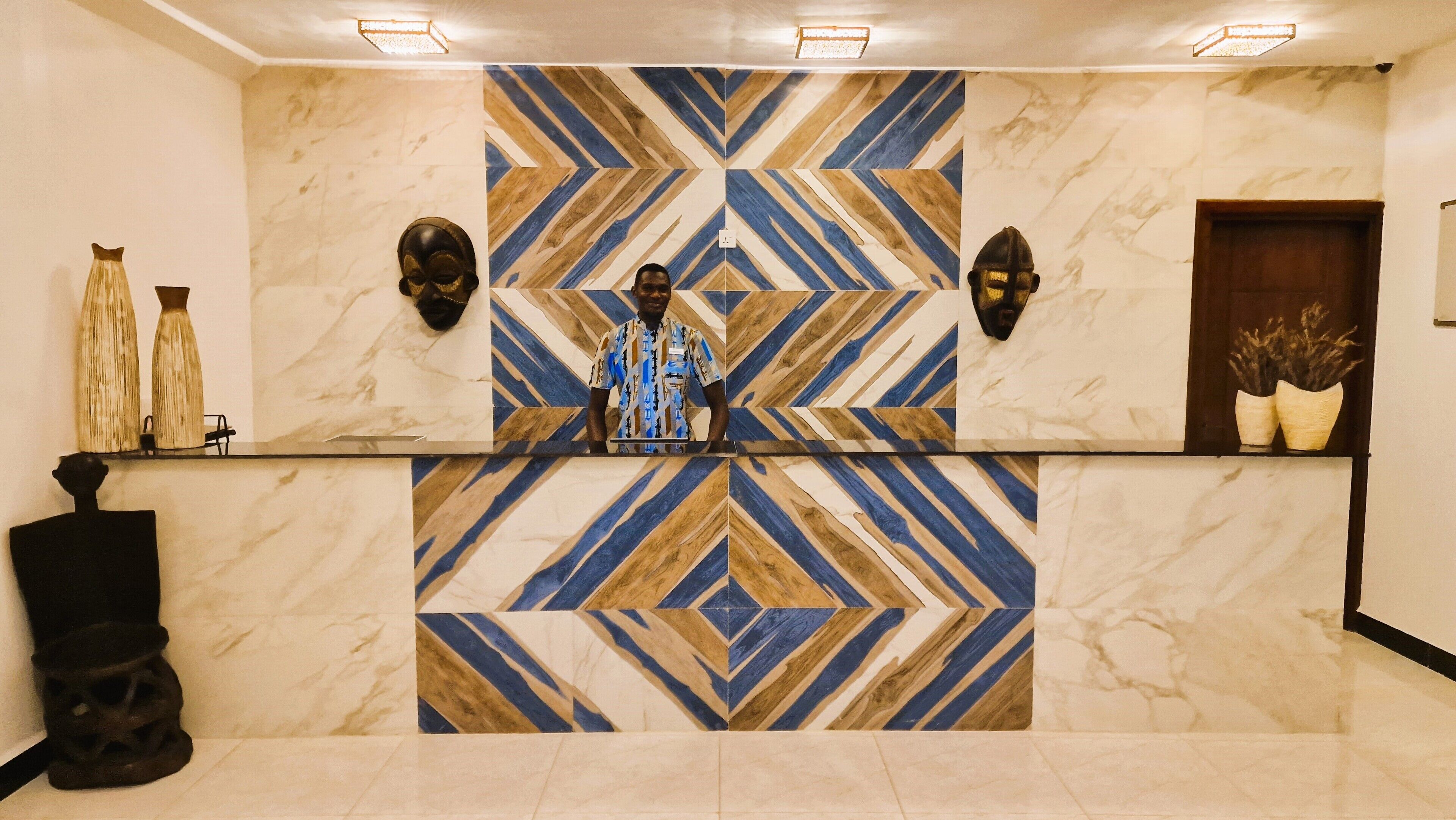A reception desk with a geometric patterned wall, two masks on either side, and a potted plant on the right.