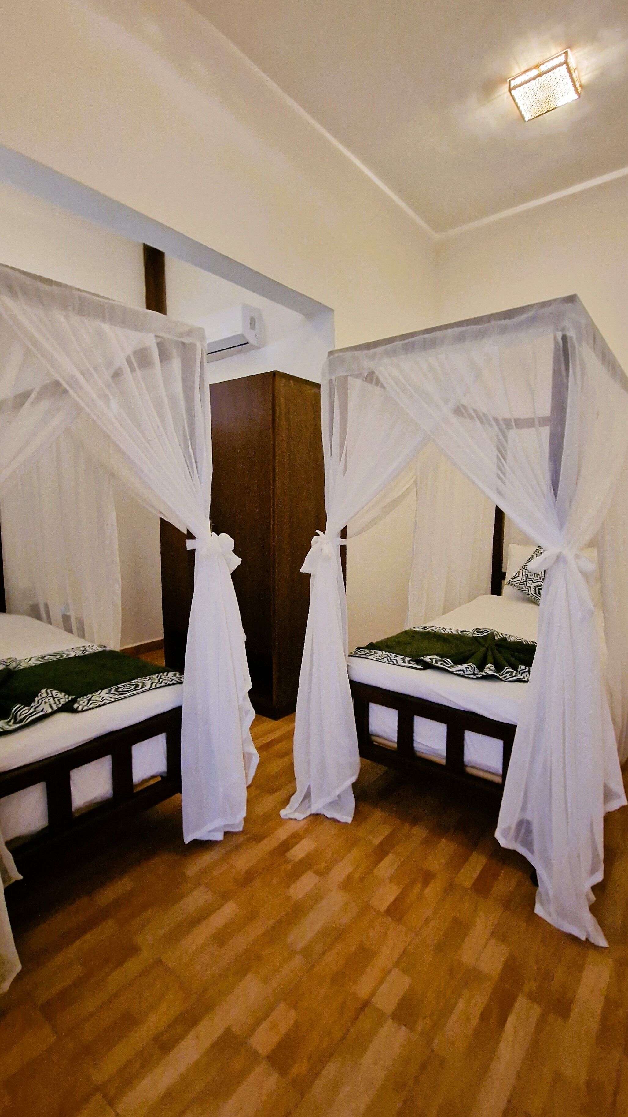 A room with two beds, each with a canopy. There is a wooden floor and a white wall with a light fixture.