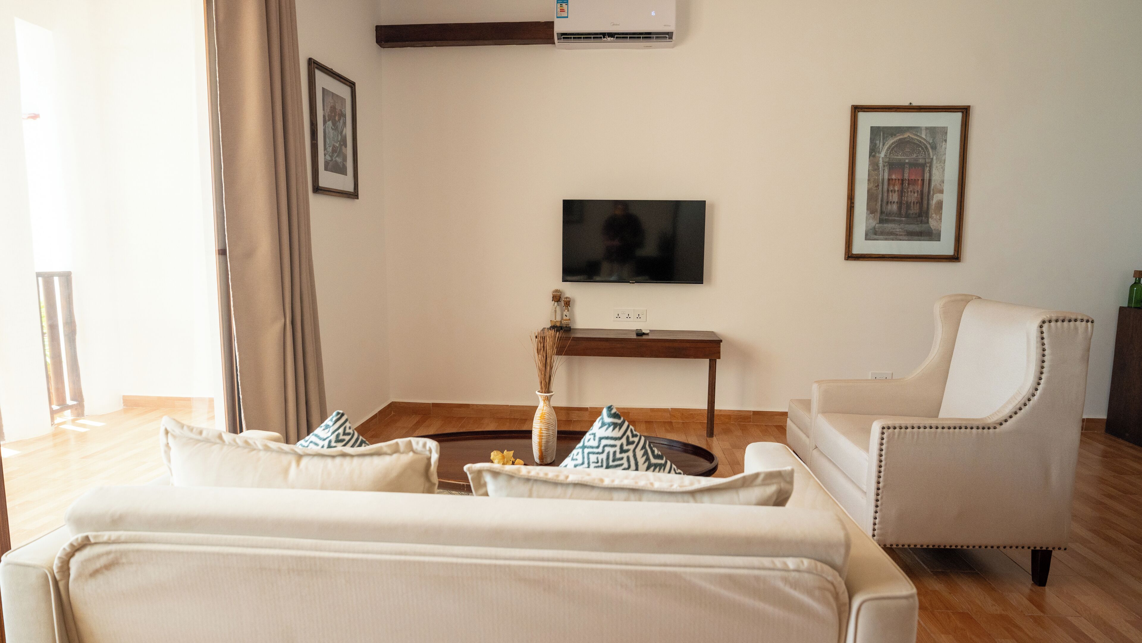 Een woonkamer met een beige bank, een bijpassende fauteuil, een flatscreen televisie, een bijzettafeltje met decoratieve objecten en een lijst met een afbeelding aan de muur.