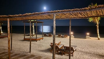 Un espacio de descanso frente a la playa con sillas de madera y una estructura de techo de paja, con vista al océano.