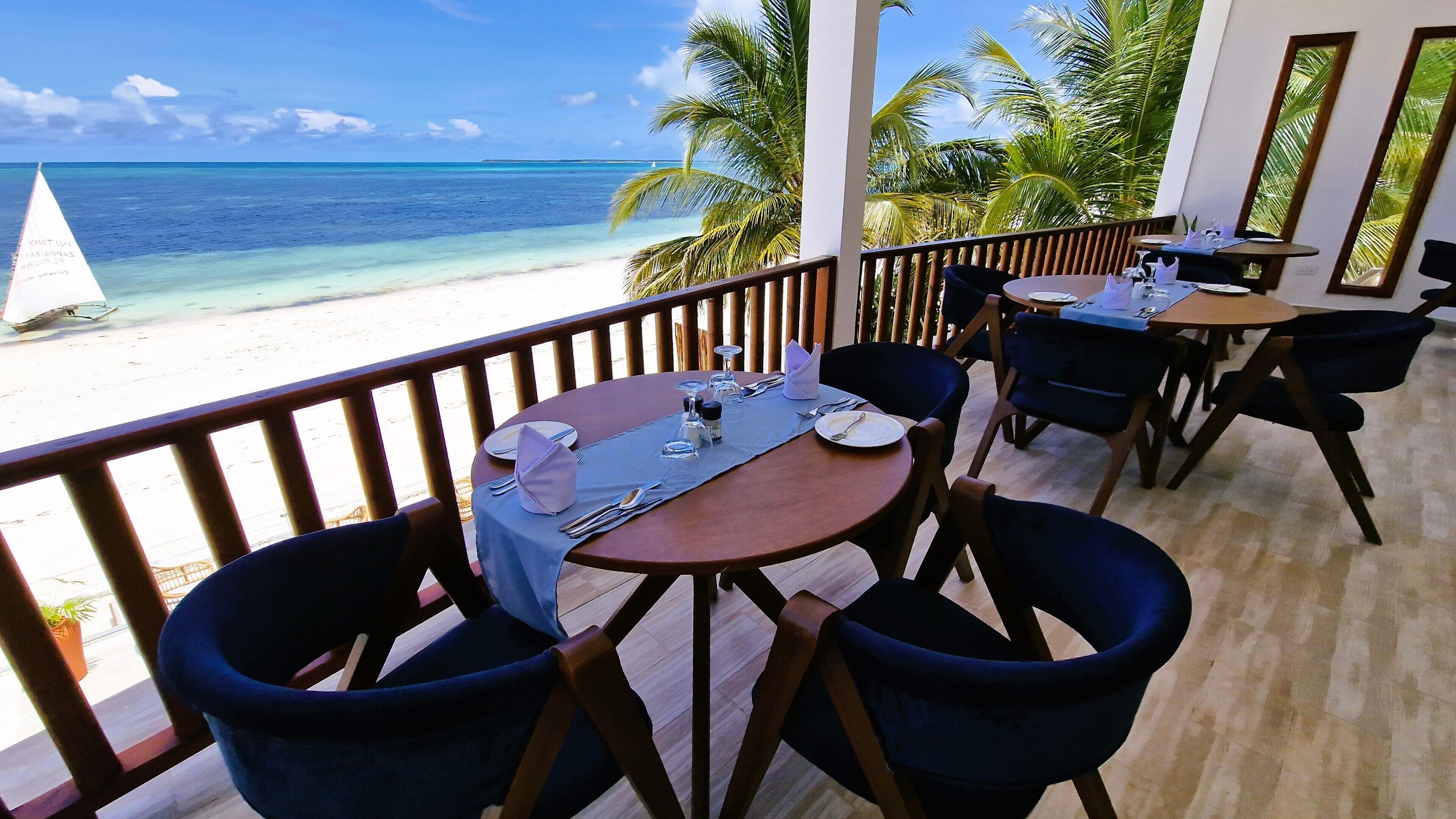 A restaurant with a view of the beach and ocean. It has wooden tables and chairs, and a blue tablecloth.