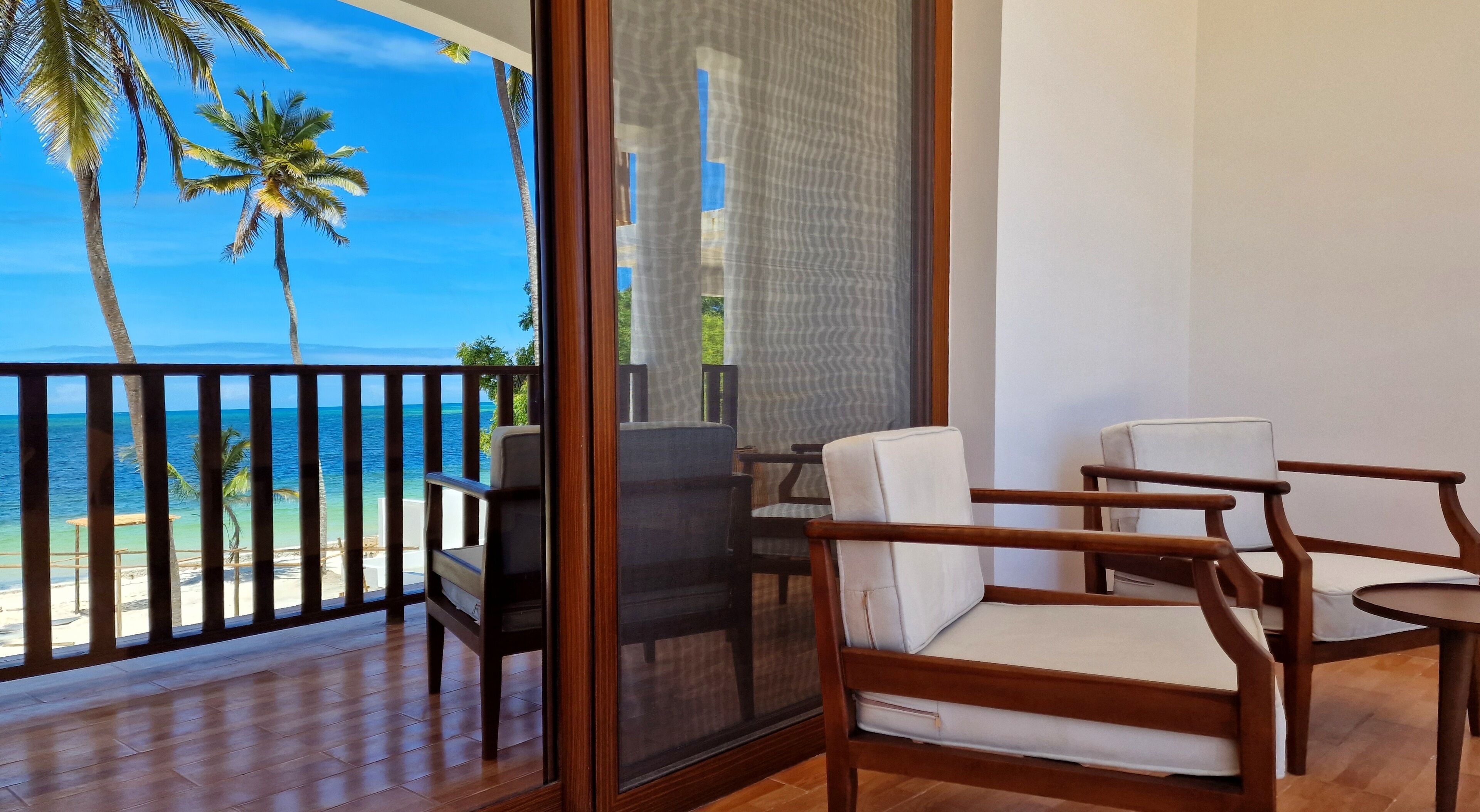 A balcony with wooden chairs and a table, overlooking a beach with palm trees and the ocean.