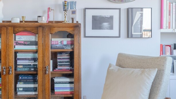 Une pièce avec une armoire en bois remplie de livres, un miroir rond et un fauteuil beige.