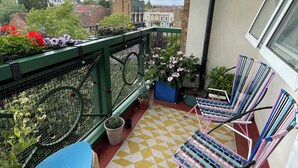 Un balcon avec un tapis à carreaux jaunes et blancs, une chaise bleue et une chaise rose. On y trouve des plantes en pot et une clôture métallique verte.