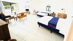 A hotel room with a large bed, a sofa, a small table, and a chair. There is a window with curtains, a small table with a plant, and a cabinet with a plant on top. - Almendra Beach Place (Los Cóbanos)
