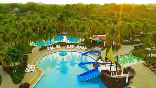A large swimming pool with a pirate ship slide, surrounded by palm trees and lounge chairs. - Almendra Beach Place (Los Cóbanos)
