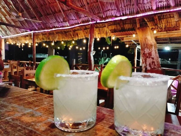 Deux verres contenant une boisson claire, décorés de rondelles de citron vert, sont posés sur une table sous un toit de chaume, illuminée par des guirlandes lumineuses.